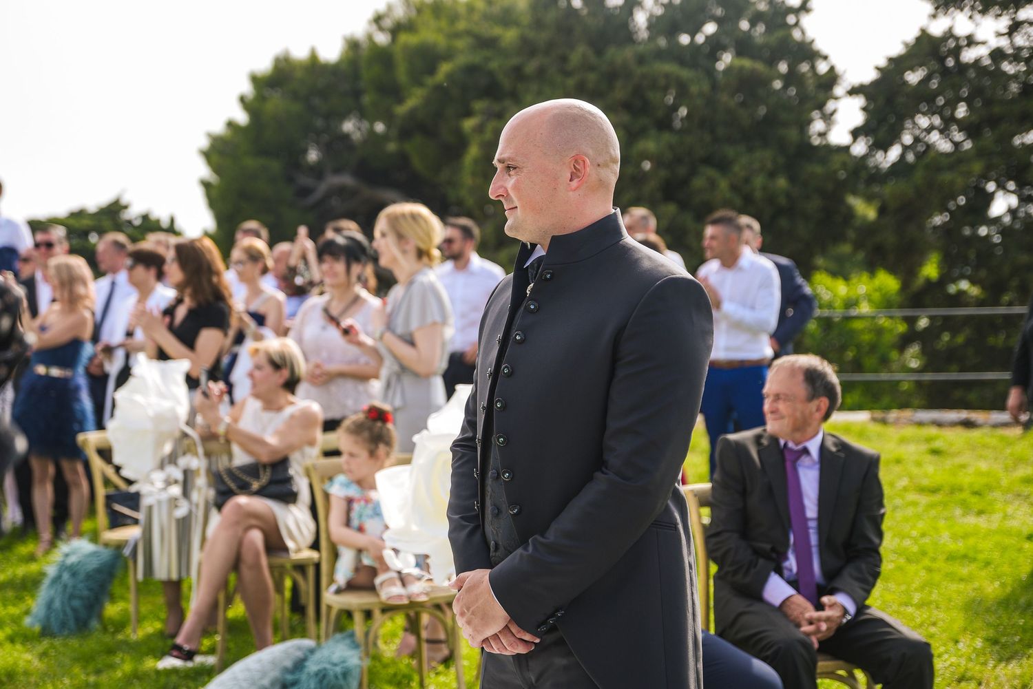 Groom in dark suit standing at outdoor wedding ceremony with seated guests.