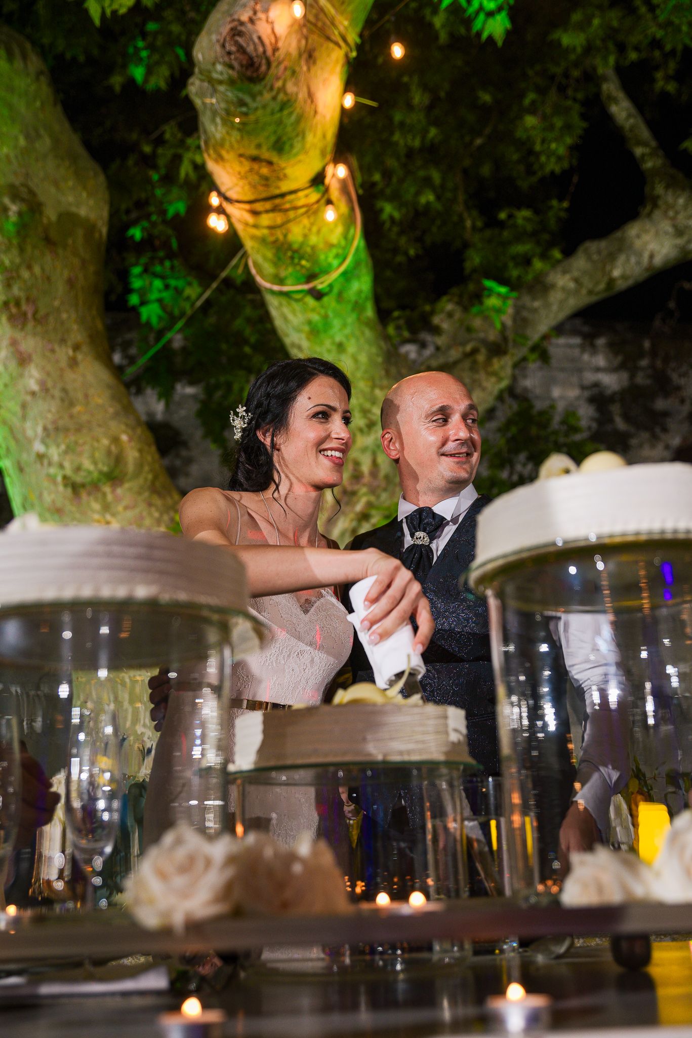 Smiling bride and groom cut their wedding cake together under a beautifully lit outdoor tree at night.