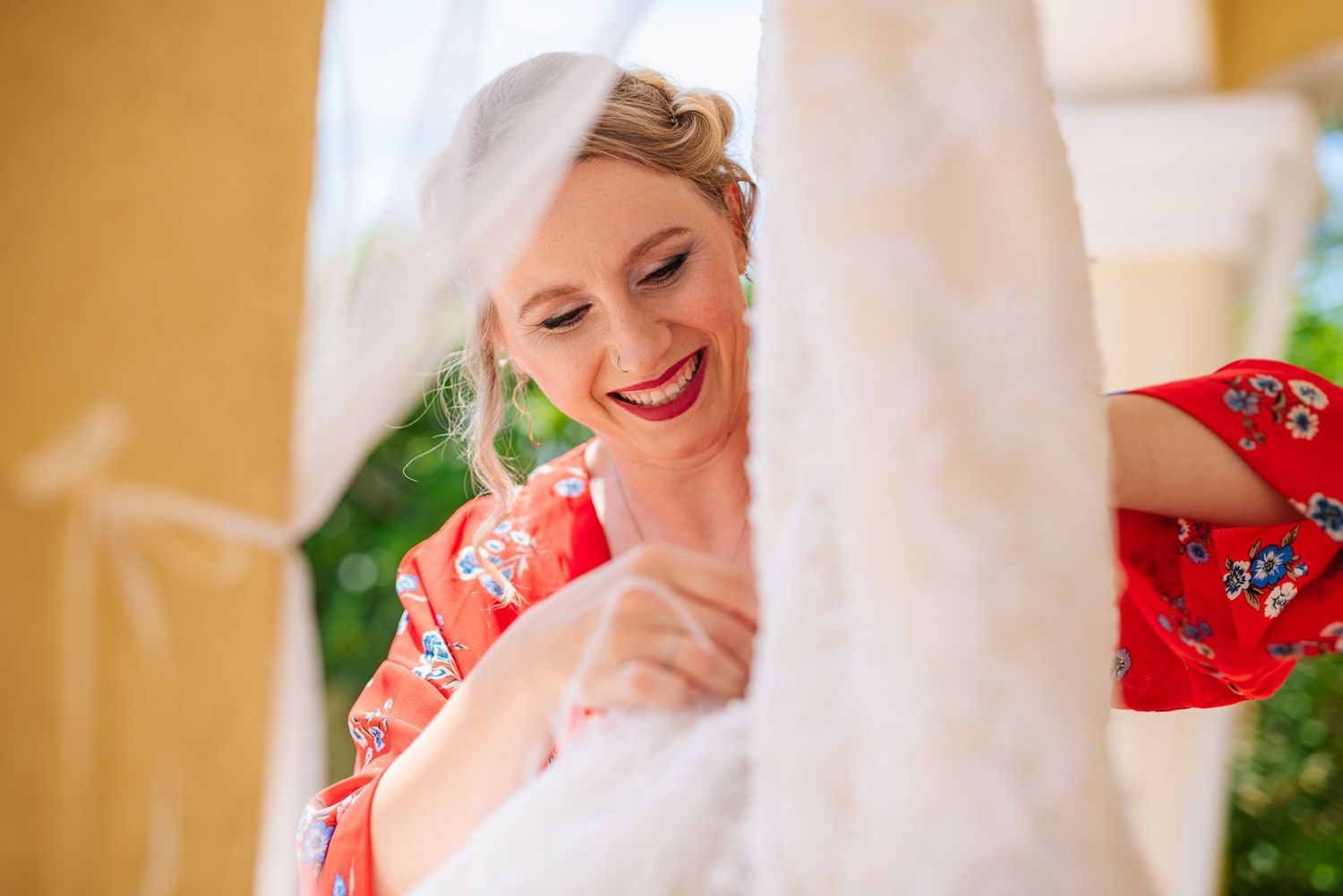 Smiling bridesmaid in red floral robe admires hanging white wedding dress indoors.