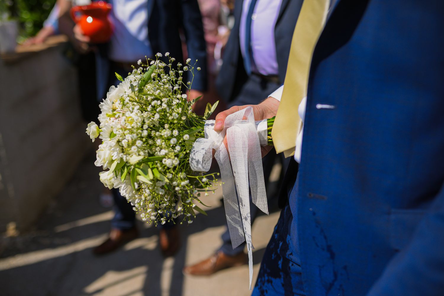 Close-up of white wedding bouquet with ribbon held by person in blue suit at outdoor ceremony.