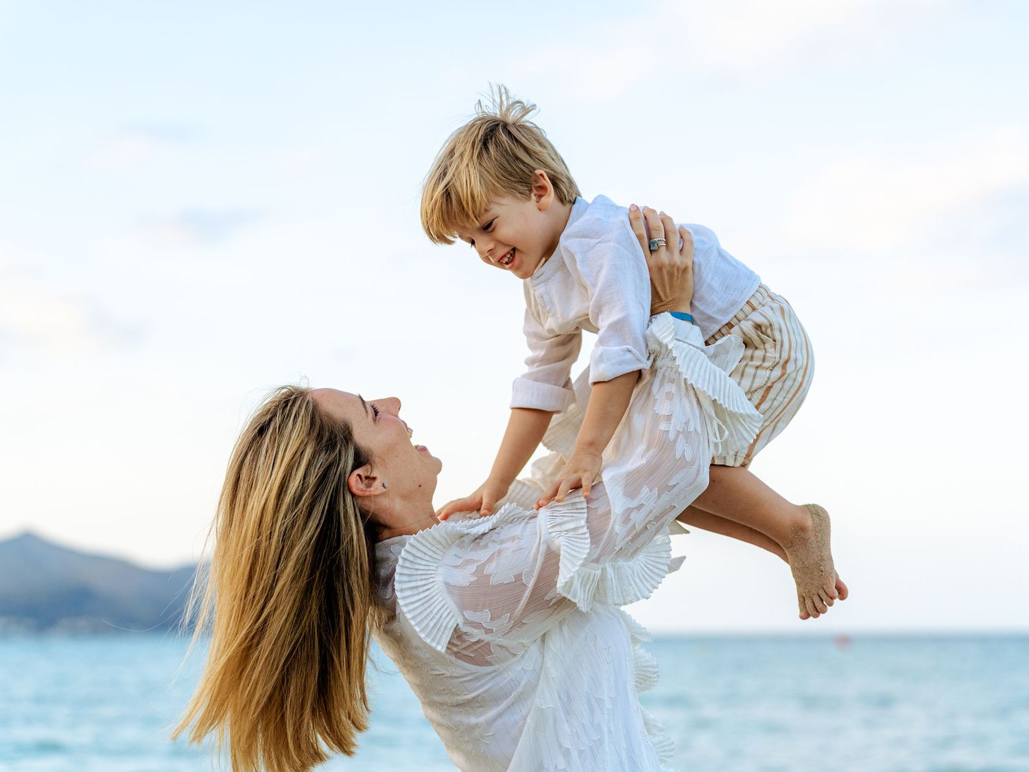 Mother in white dress joyfully lifting toddler boy at tropical beach.