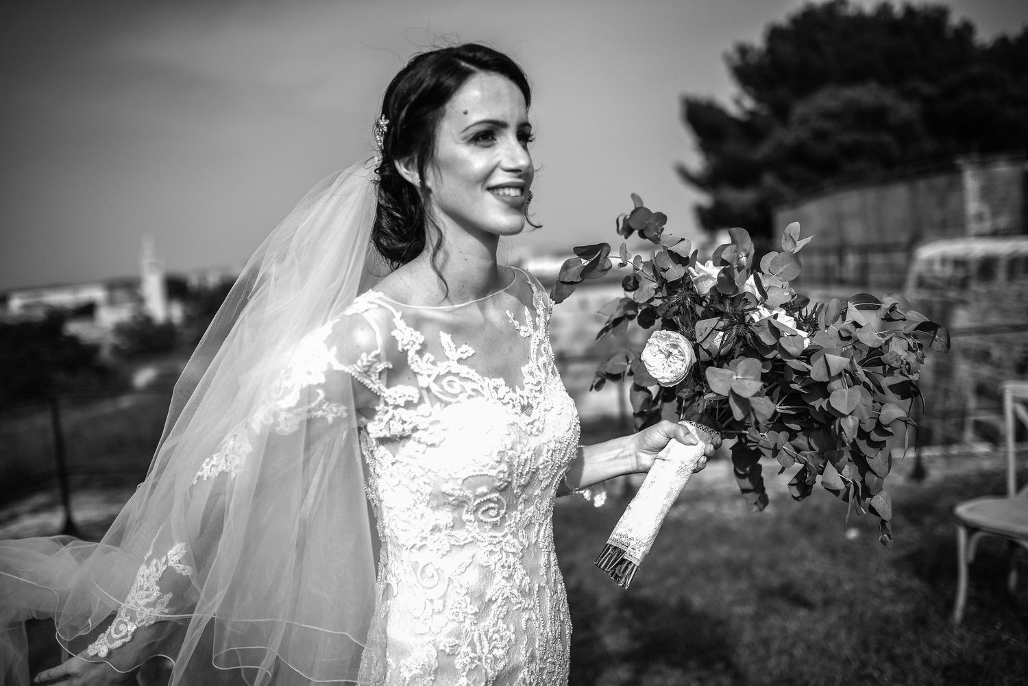 Elegant bride in lace wedding gown holding a bouquet, black and white portrait outdoors.