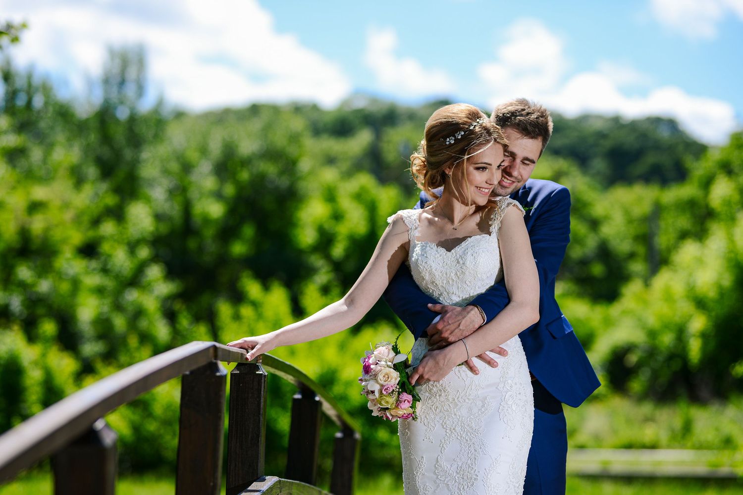 Groom in blue suit embraces bride in lace wedding dress on a wooden bridge surrounded by lush green scenery.