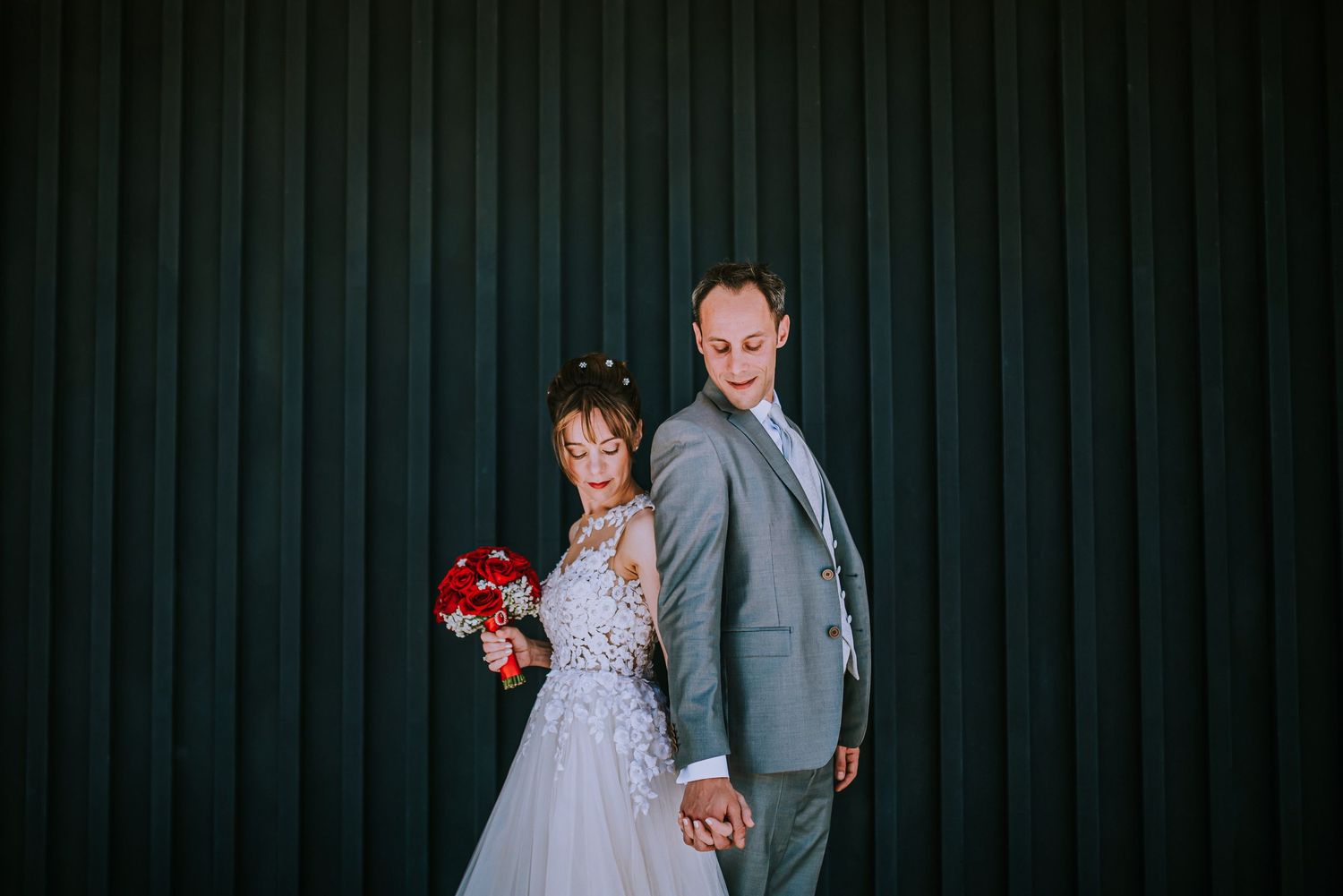 Bride and groom pose back-to-back holding hands with a red bouquet against a dark wall.