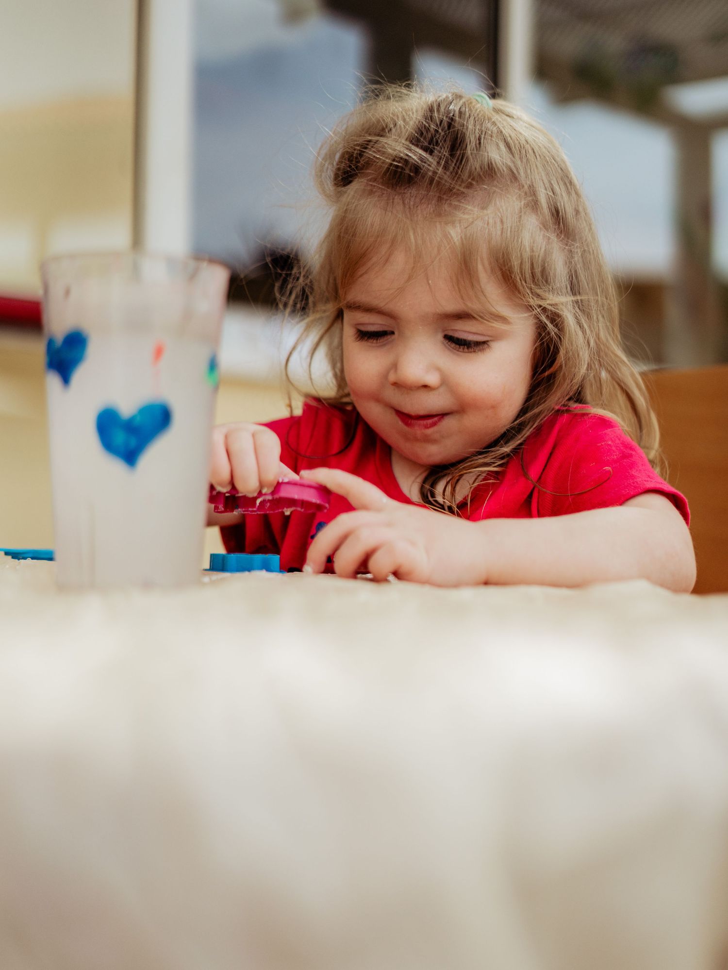 Toddler girl in pink shirt playing with colorful playdough near a decorated cup with blue hearts.