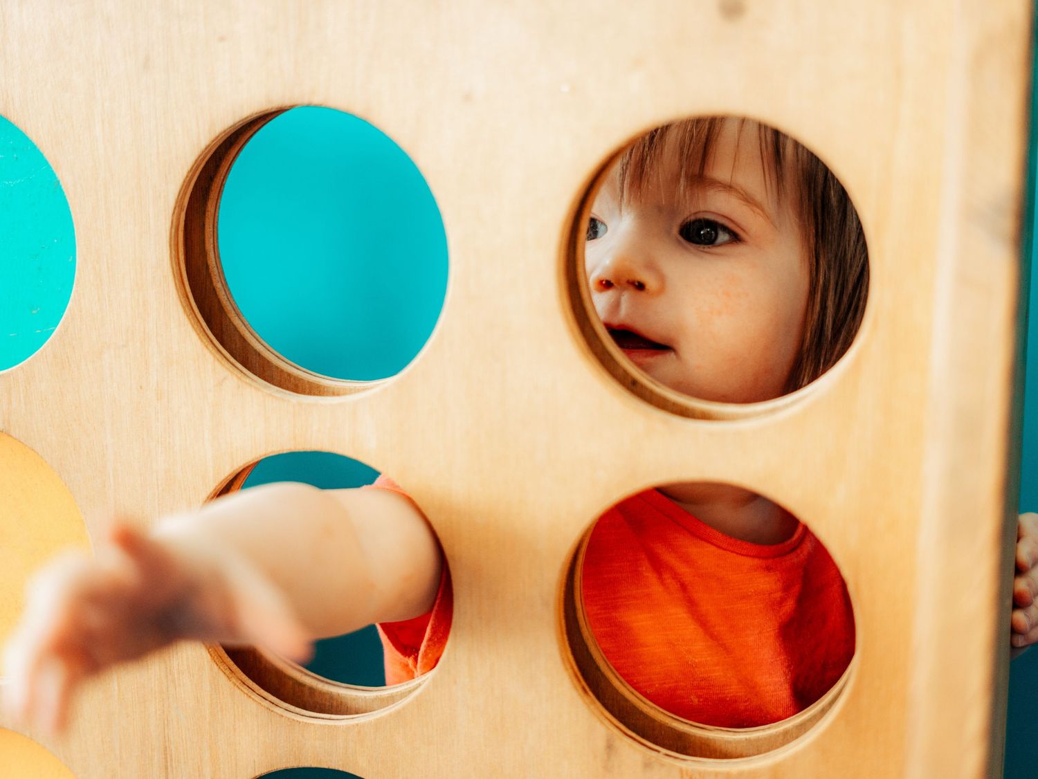 Curious toddler girl in orange shirt looking through colorful circular holes in wooden play wall.