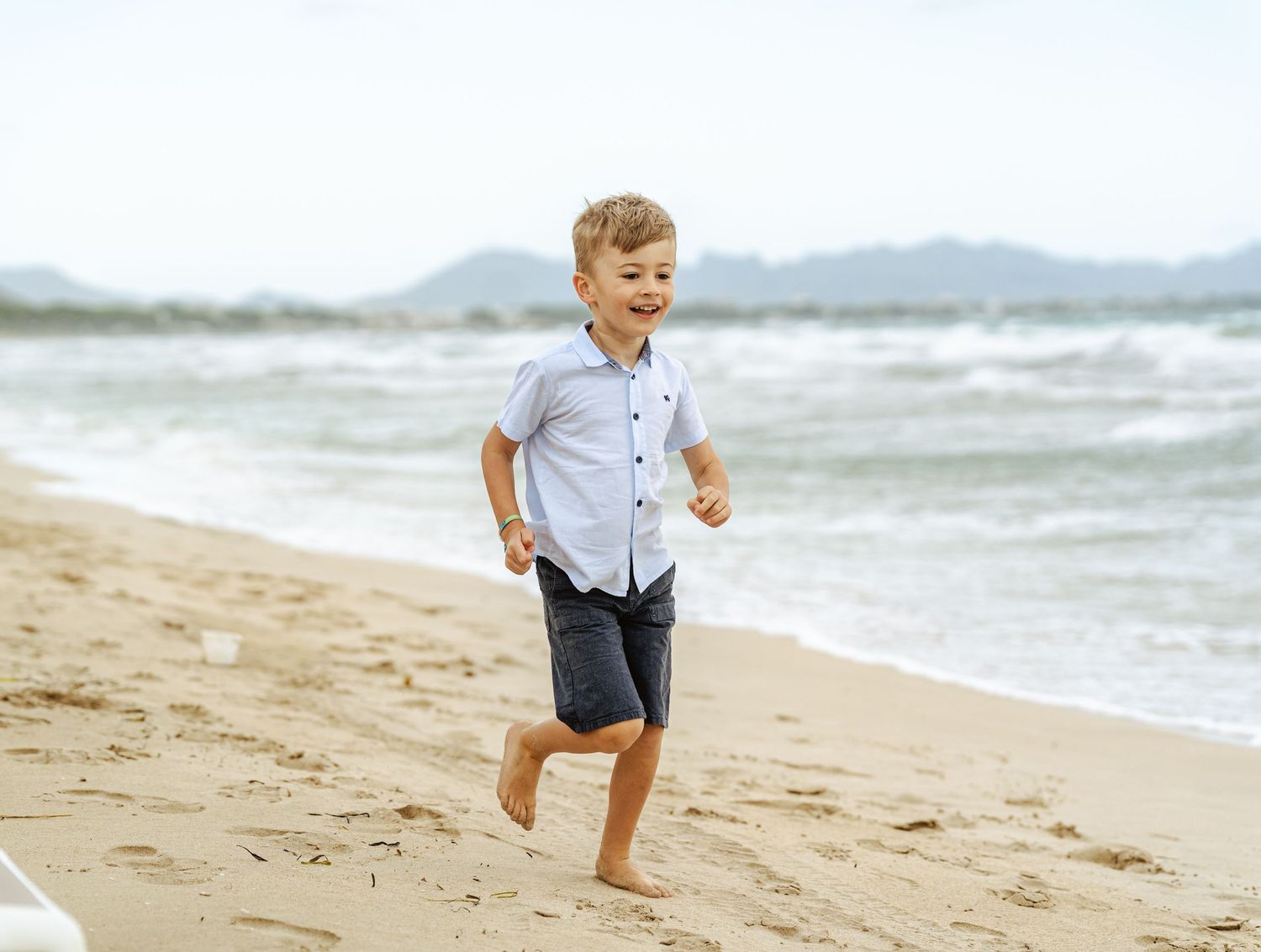 Happy young boy running barefoot along sandy beach with ocean waves behind.