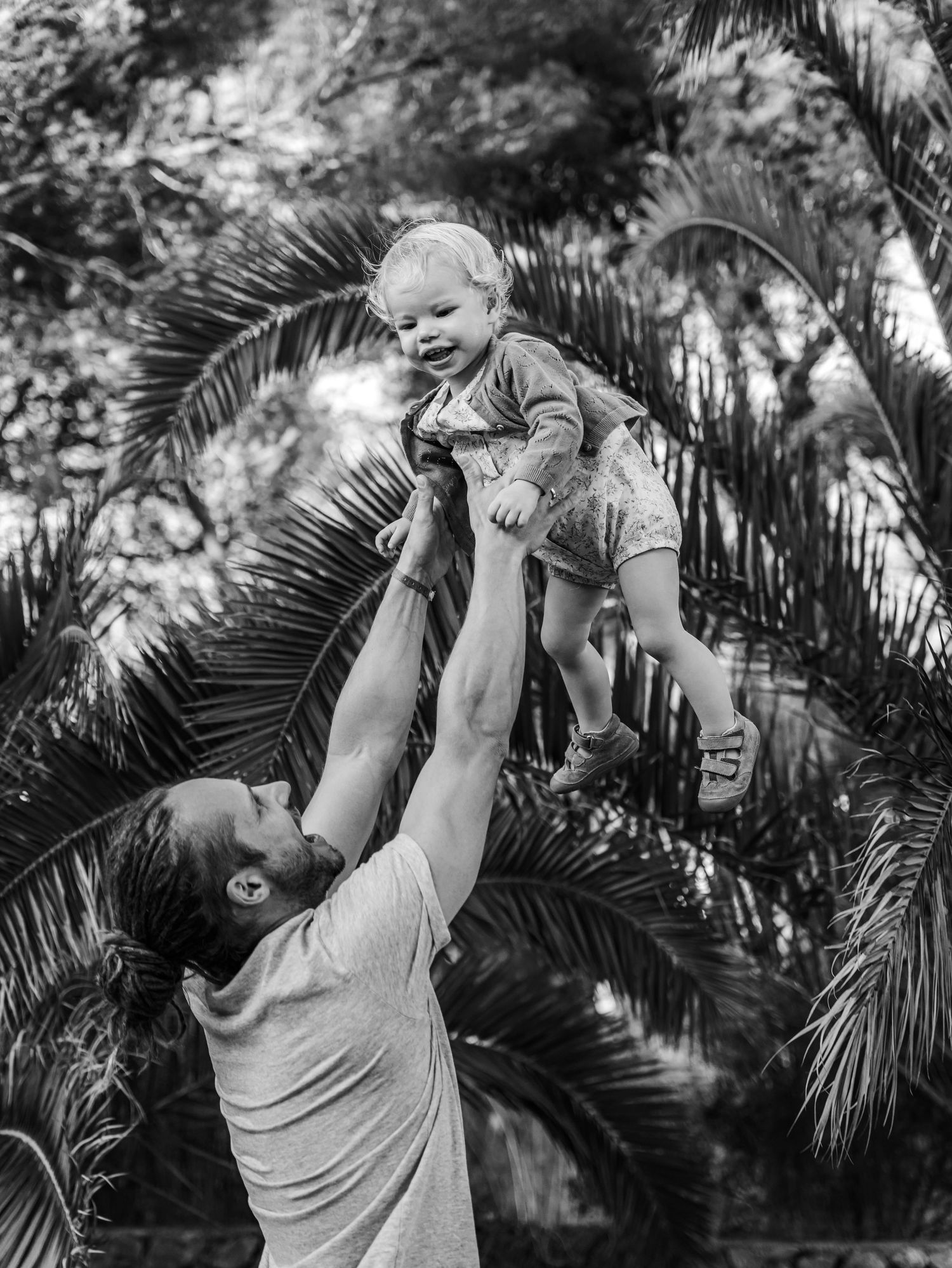 Black and white photo of father lifting laughing baby high in the air among tropical palm trees.