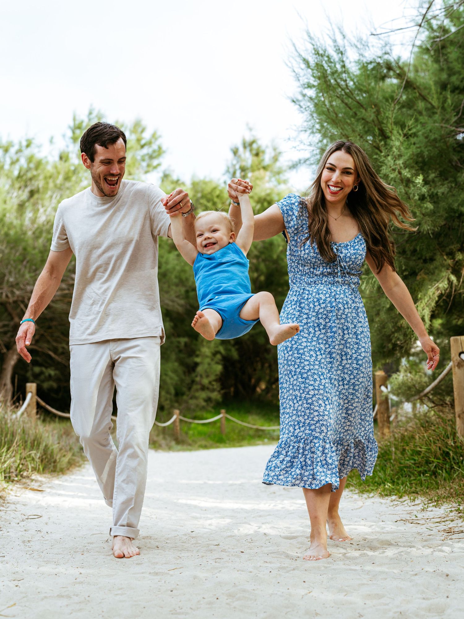 Happy family swinging baby on sandy beach path surrounded by lush green trees on sunny day.