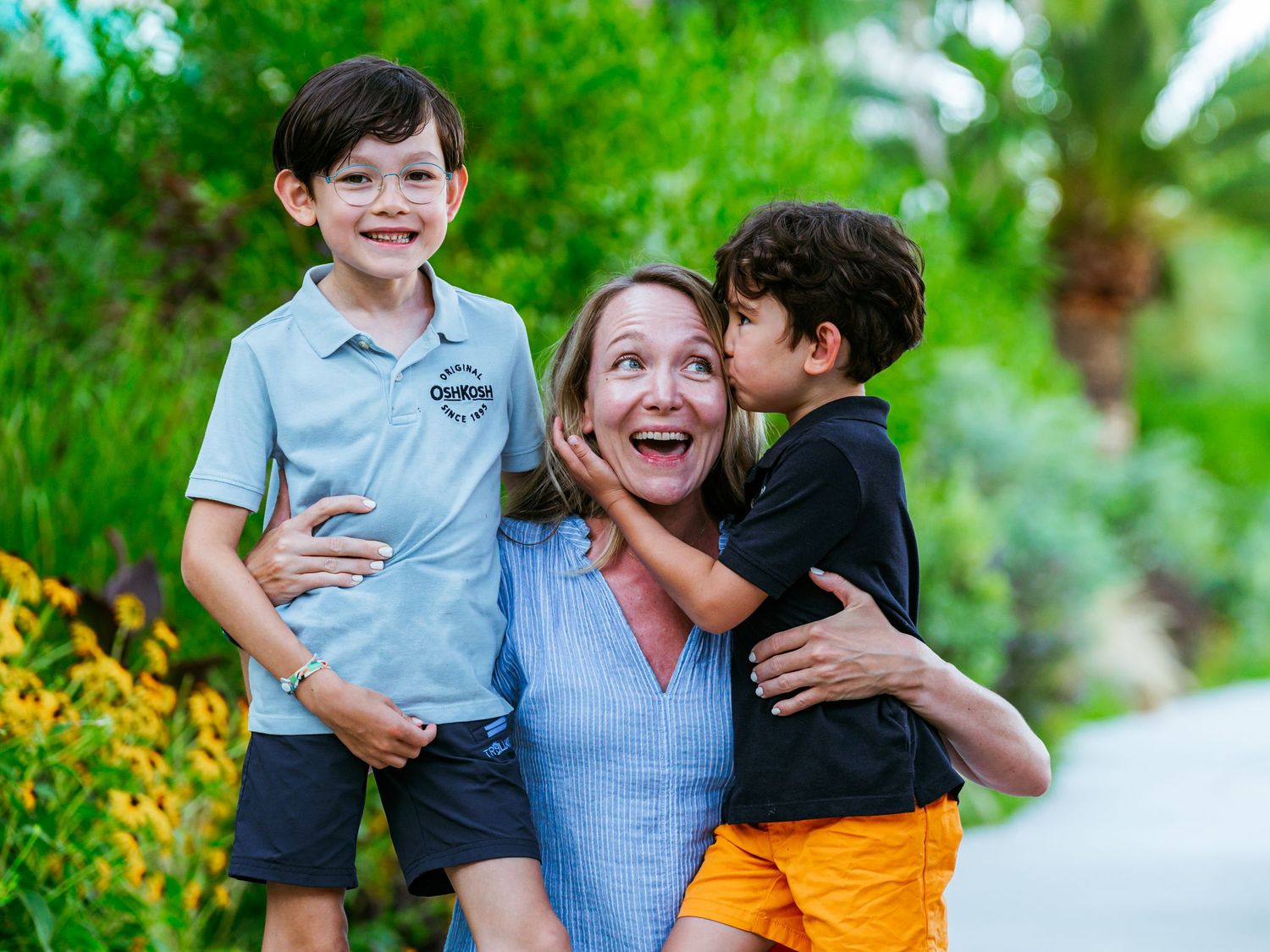 Joyful mother laughing as two young sons hug and kiss her cheeks outdoors.