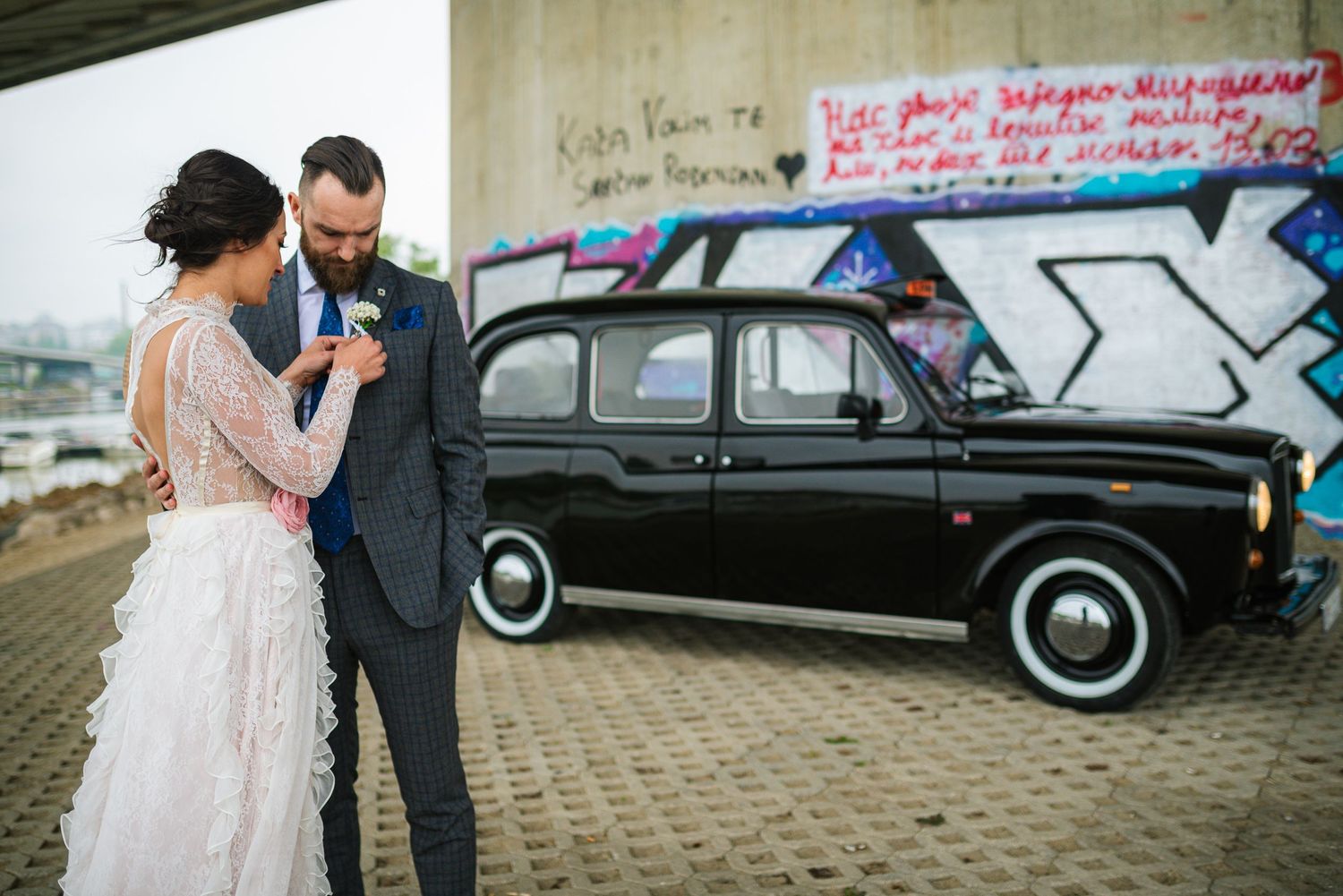 Bride and groom standing before black London taxi cab with colorful graffiti wall backdrop on wedding day.