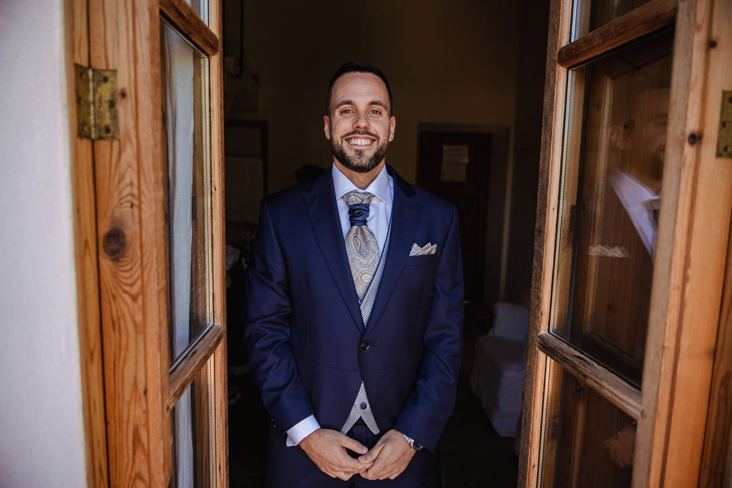 Smiling groom in a navy blue suit standing in a wooden doorway on his wedding day.