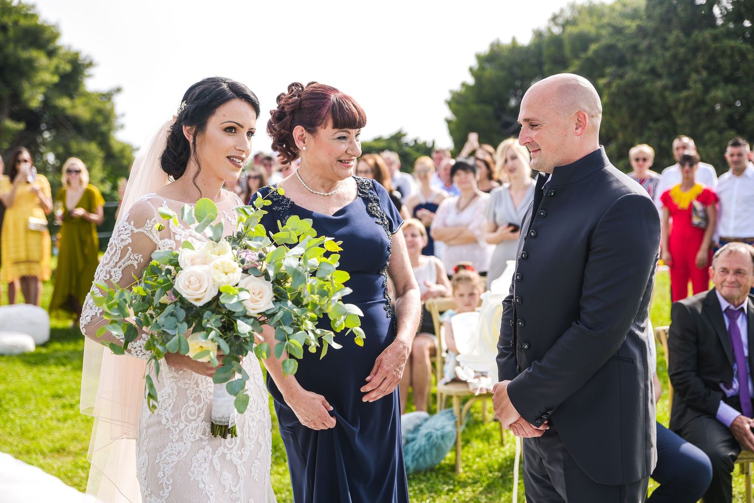 Bride smiling with bouquet meets groom at outdoor wedding ceremony on green lawn.
