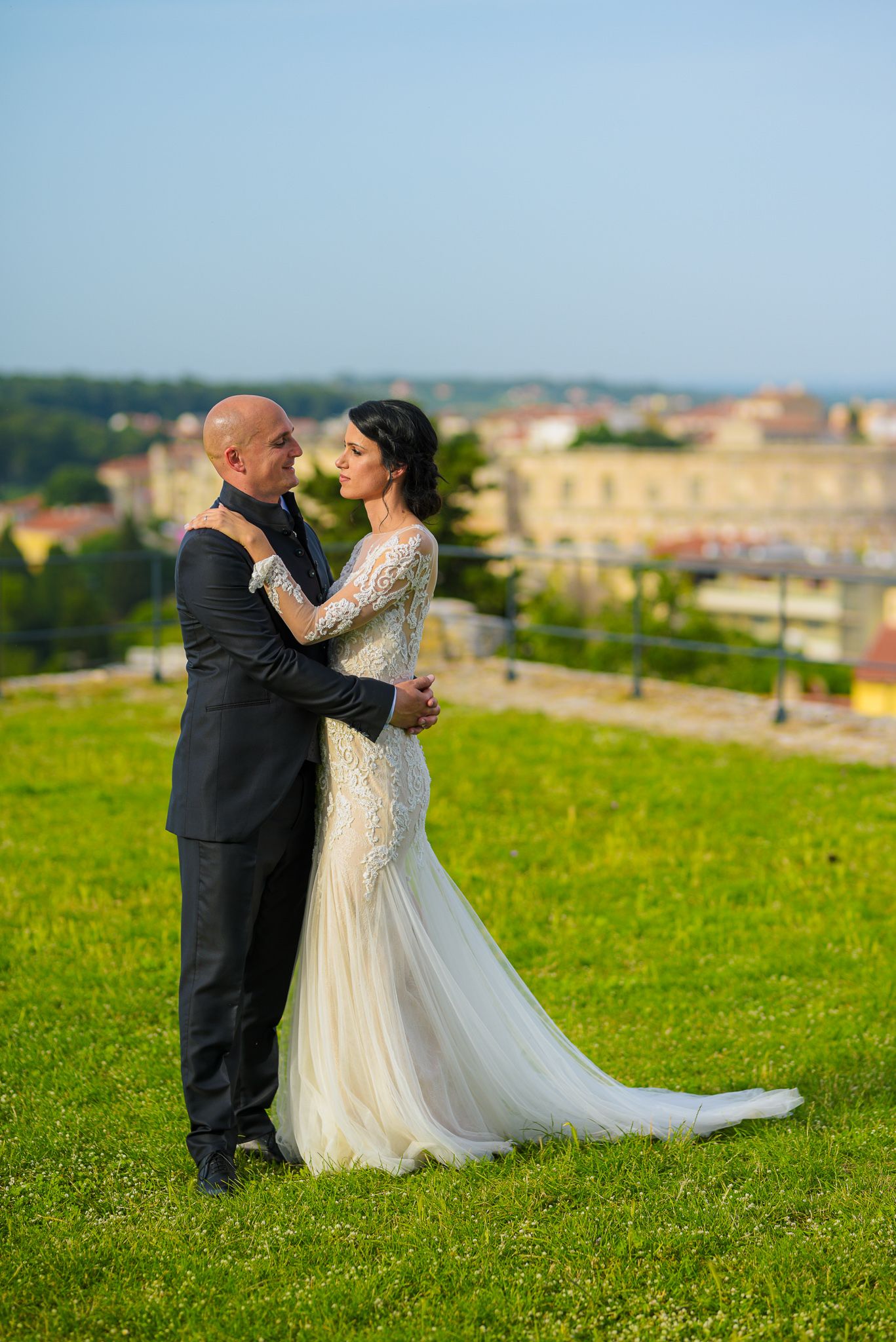 Bride and groom embracing on a green lawn with a panoramic town view behind them.