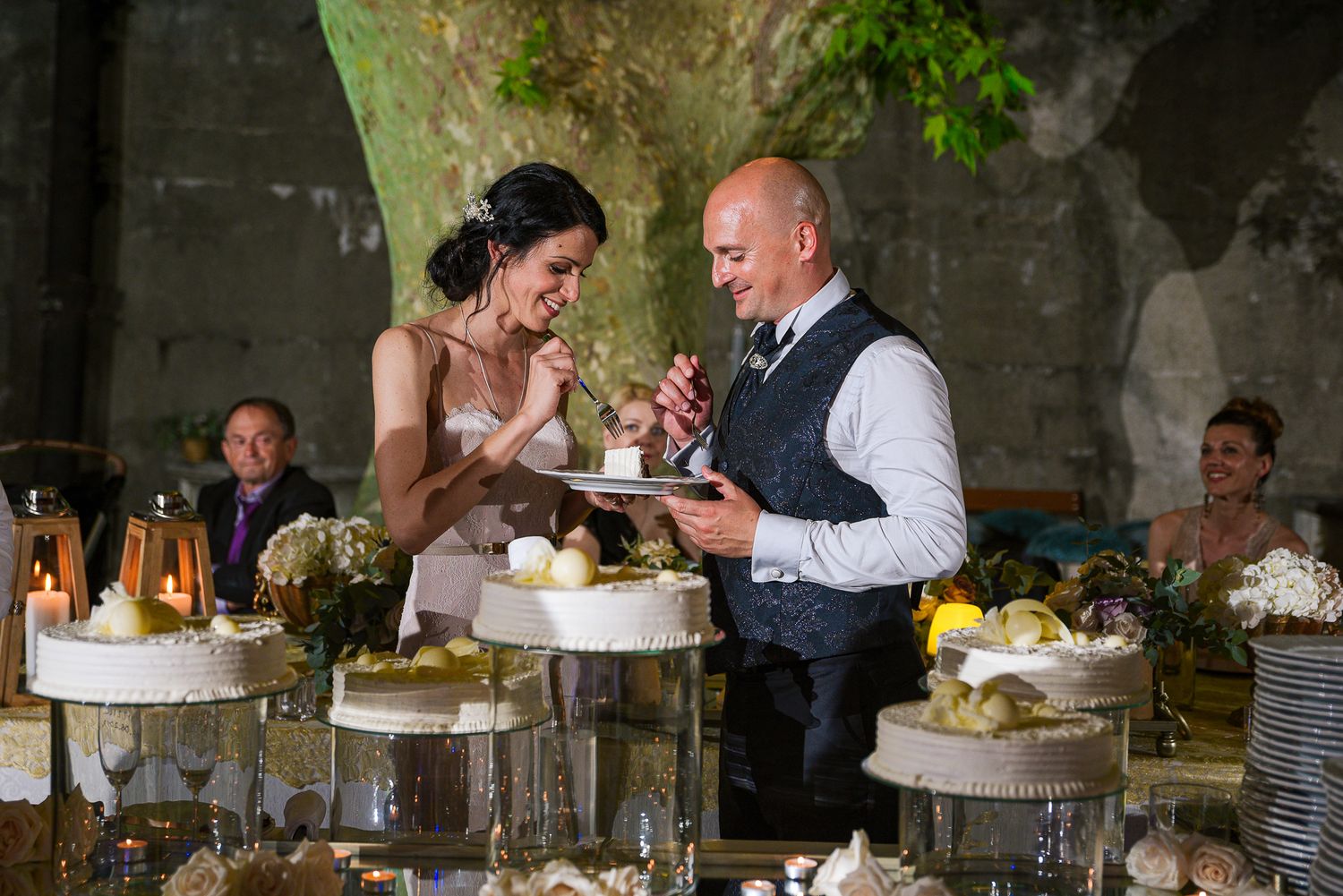 Bride and groom taste wedding cake together while guests look on at their elegant outdoor reception.