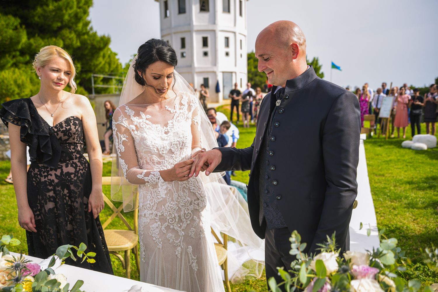 Bride and groom exchanging rings at outdoor wedding ceremony near white lighthouse with guests watching.