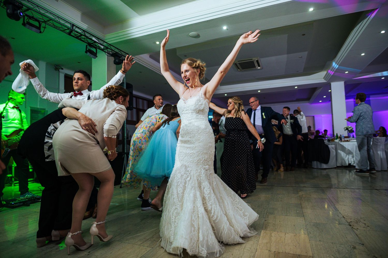 Joyful bride dancing with raised arms surrounded by guests at wedding reception.