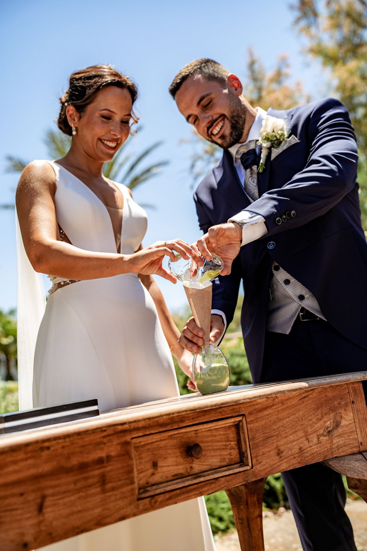Smiling bride and groom perform sand ceremony at rustic wooden table during sunny outdoor wedding.