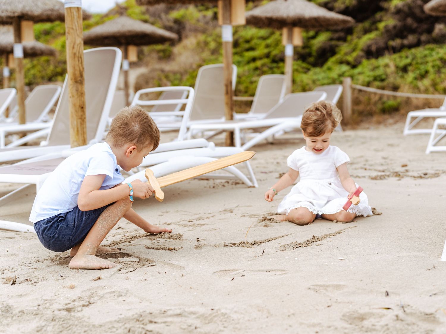 Two young children play together in the sand near beach loungers under thatched umbrellas.
