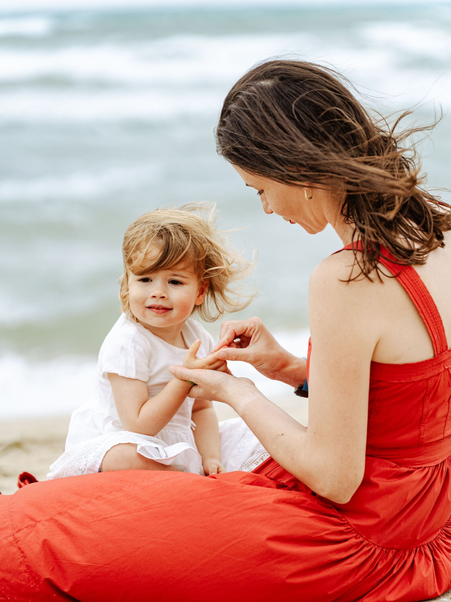 Mother in red dress sitting on beach examining something with her curly-haired toddler.