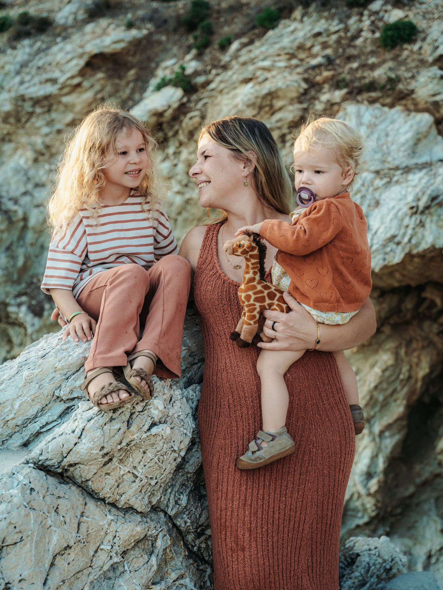 A smiling mother in a brown dress holds two young children on rocks, one clutching a giraffe toy.