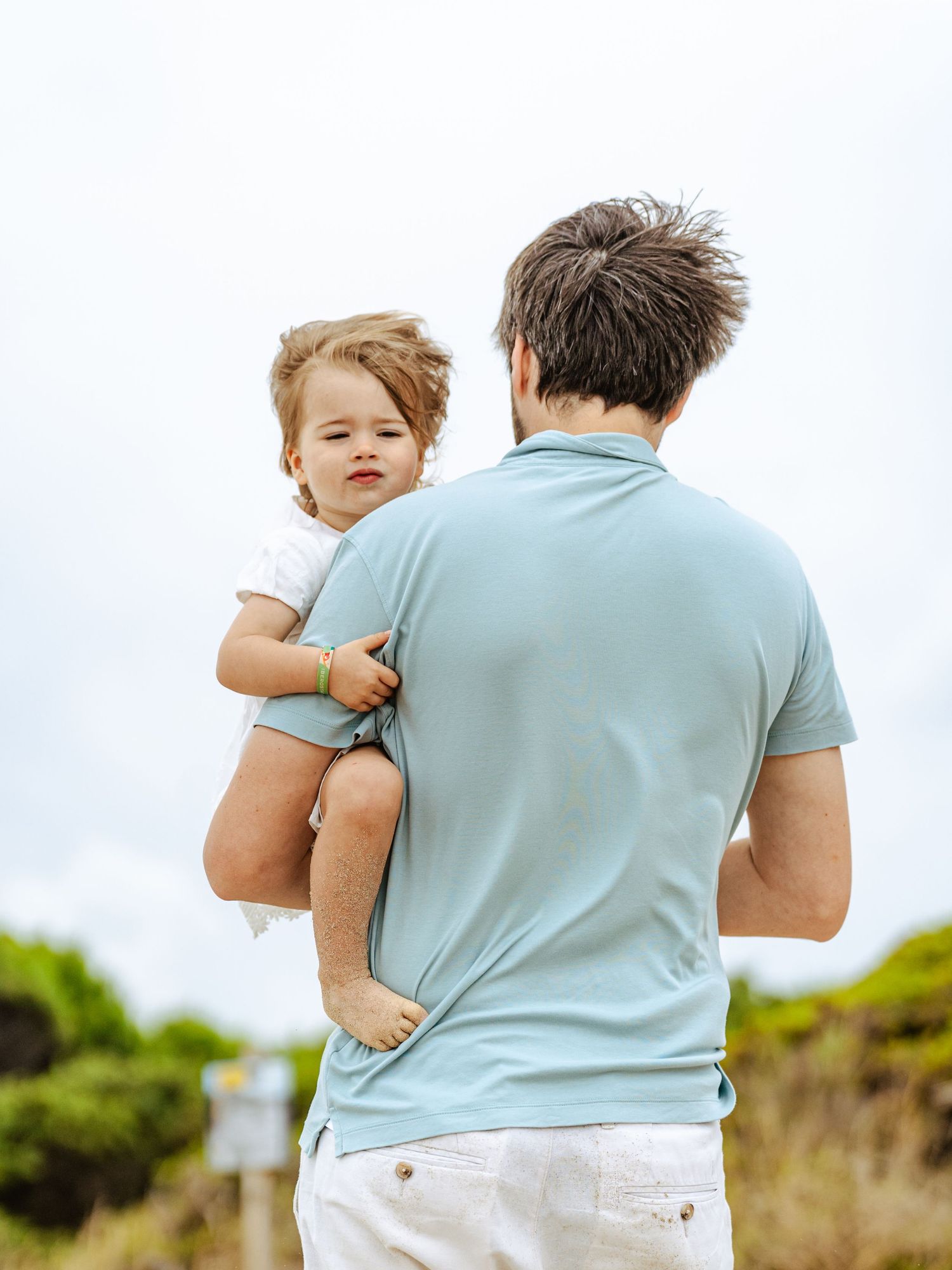 Father carrying toddler in white outfit, viewed from behind on a cloudy beach day.