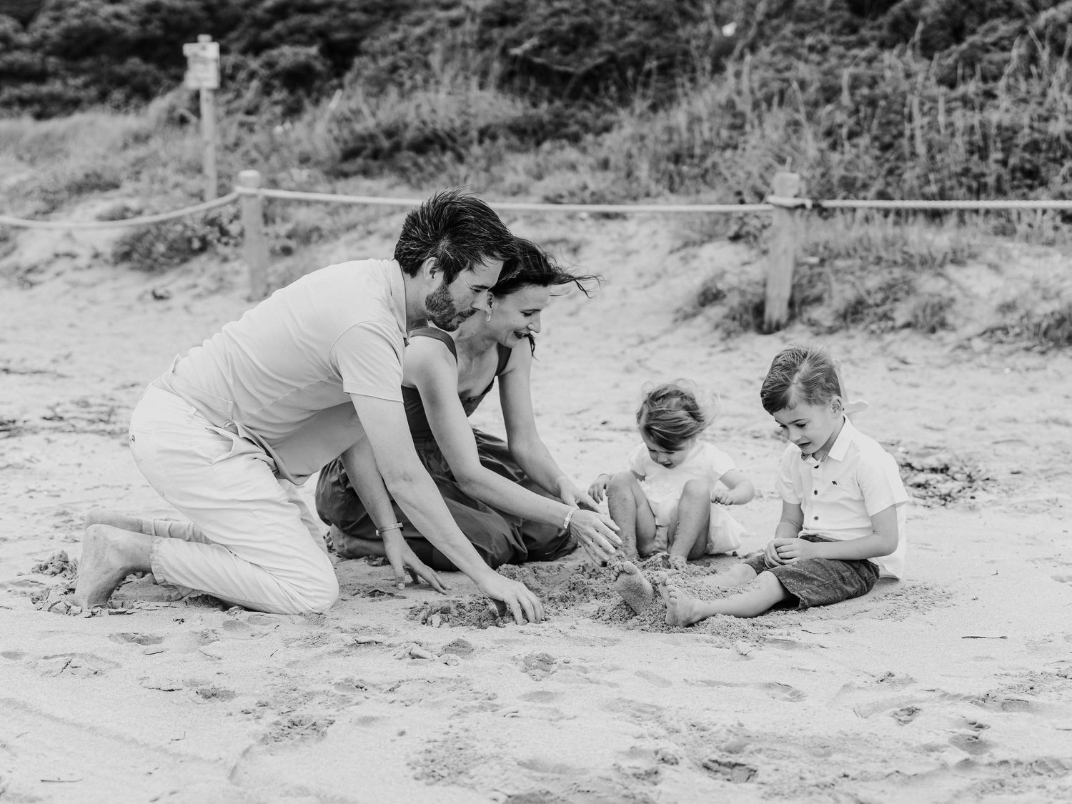 A family with two young children plays together in the sand at the beach in a black and white photo.