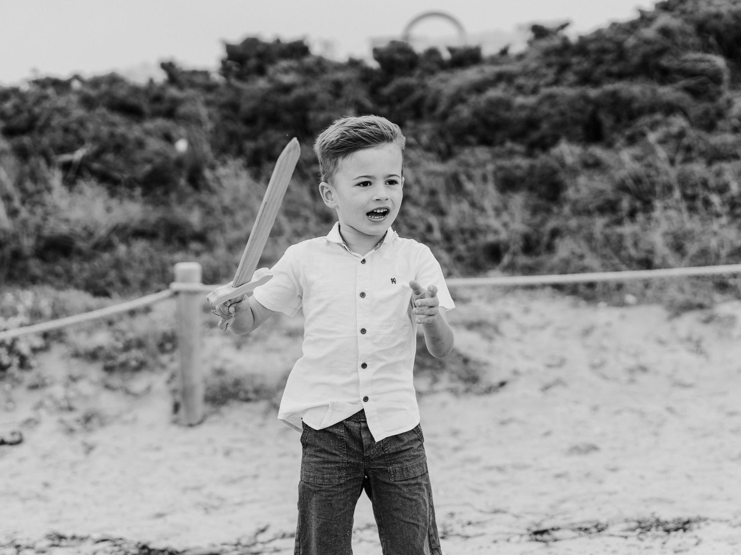 Black and white photo of young boy in white shirt wielding wooden toy sword on beach.