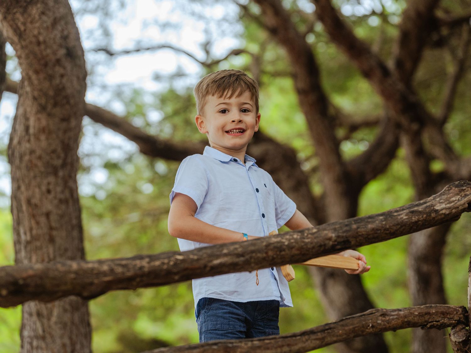 Smiling boy in light blue shirt leaning on a rustic wooden fence surrounded by lush green trees.