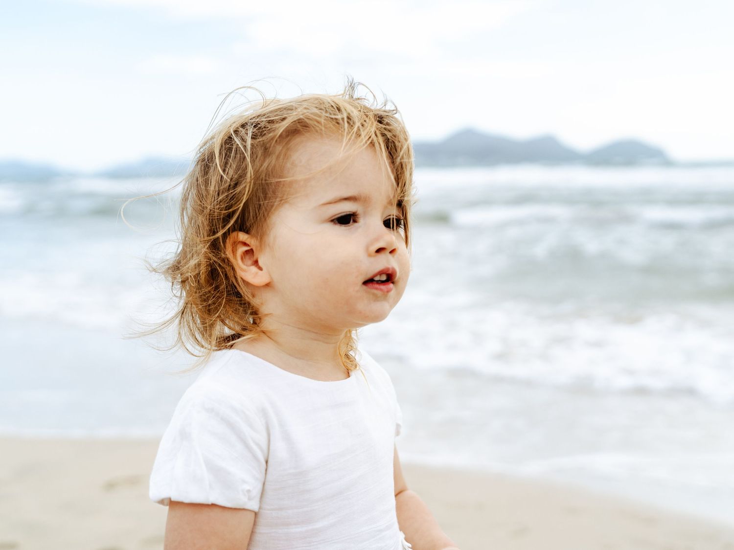Toddler girl with curly blonde hair wearing a white t-shirt standing on a sandy beach with ocean waves.
