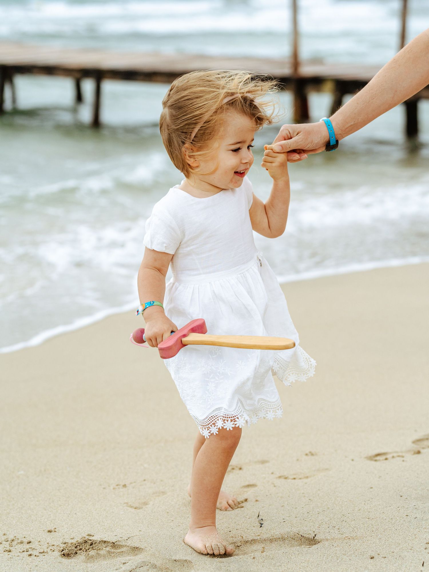 Toddler girl in white dress holding toy on sandy beach, guided by adult hand near ocean waves.