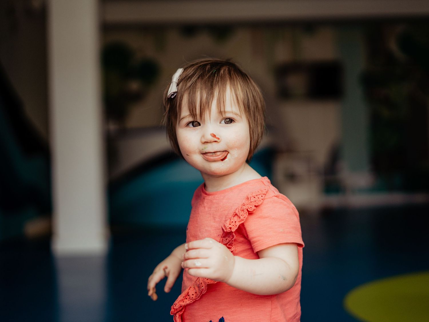 Cute toddler girl in coral ruffled top with hair bow smiling playfully indoors.