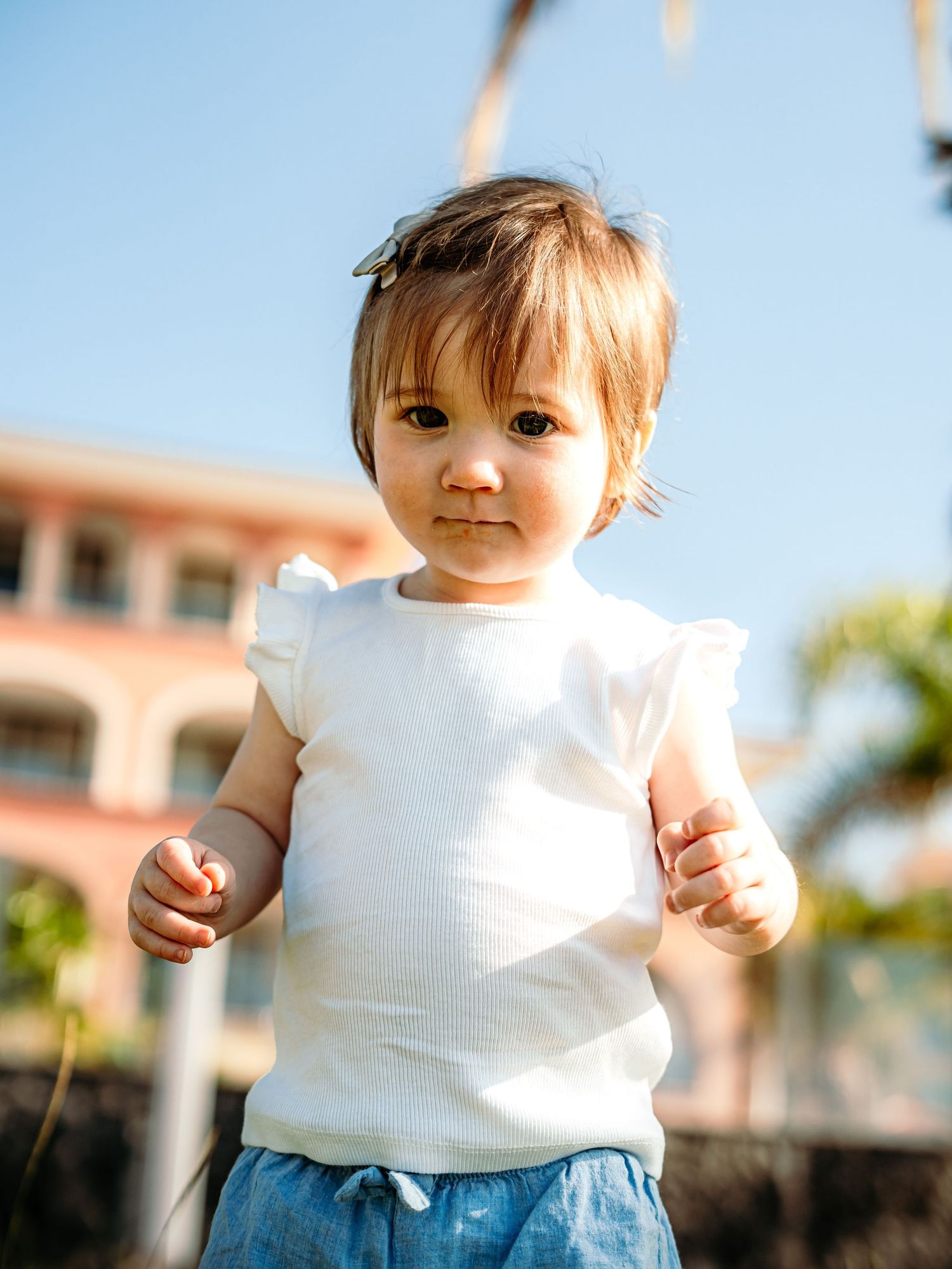 Close-up portrait of an adorable baby girl in a white ruffled top standing outdoors on a sunny day.