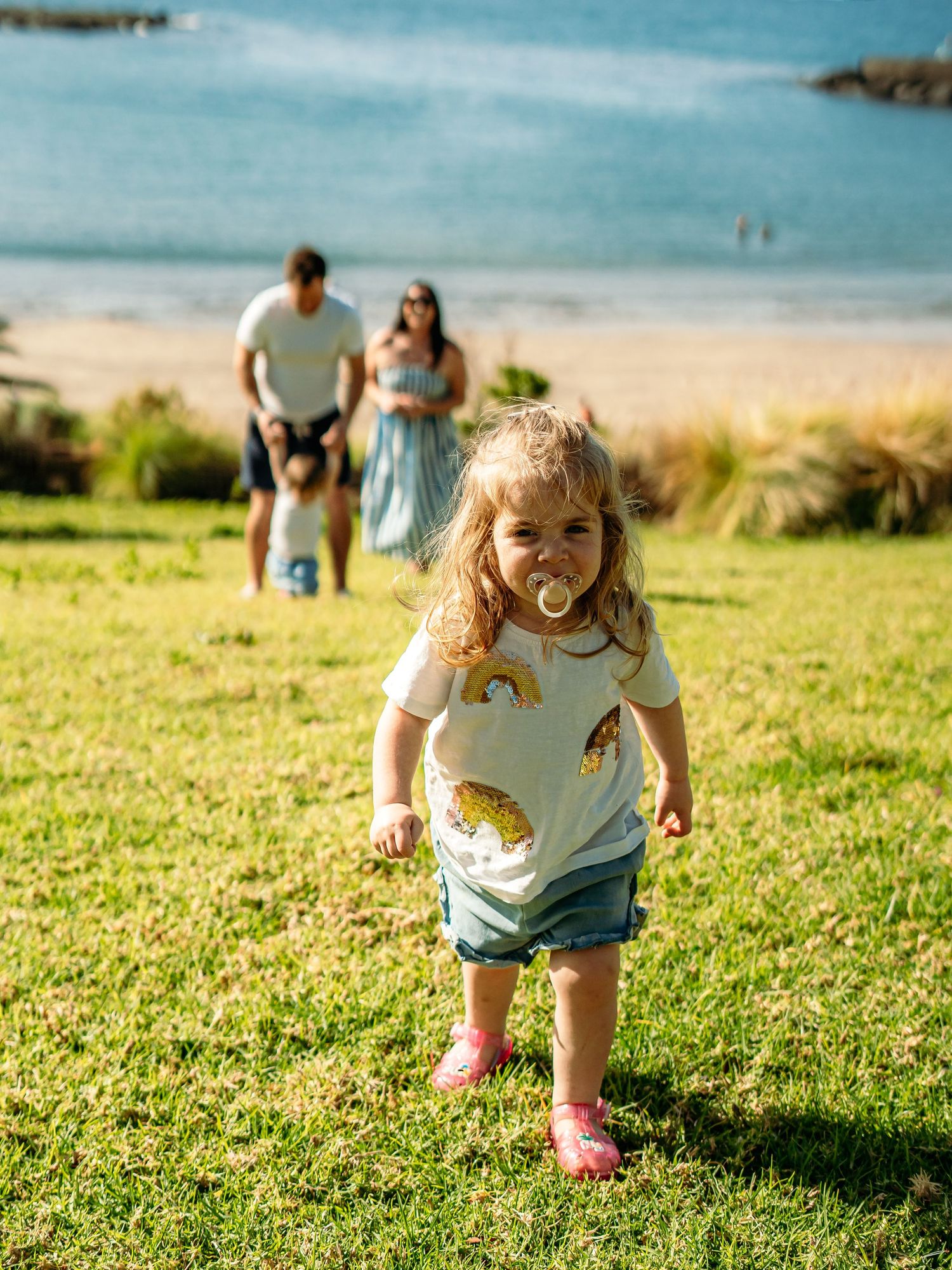 Toddler girl with pacifier walking on grass near beach with family in background.