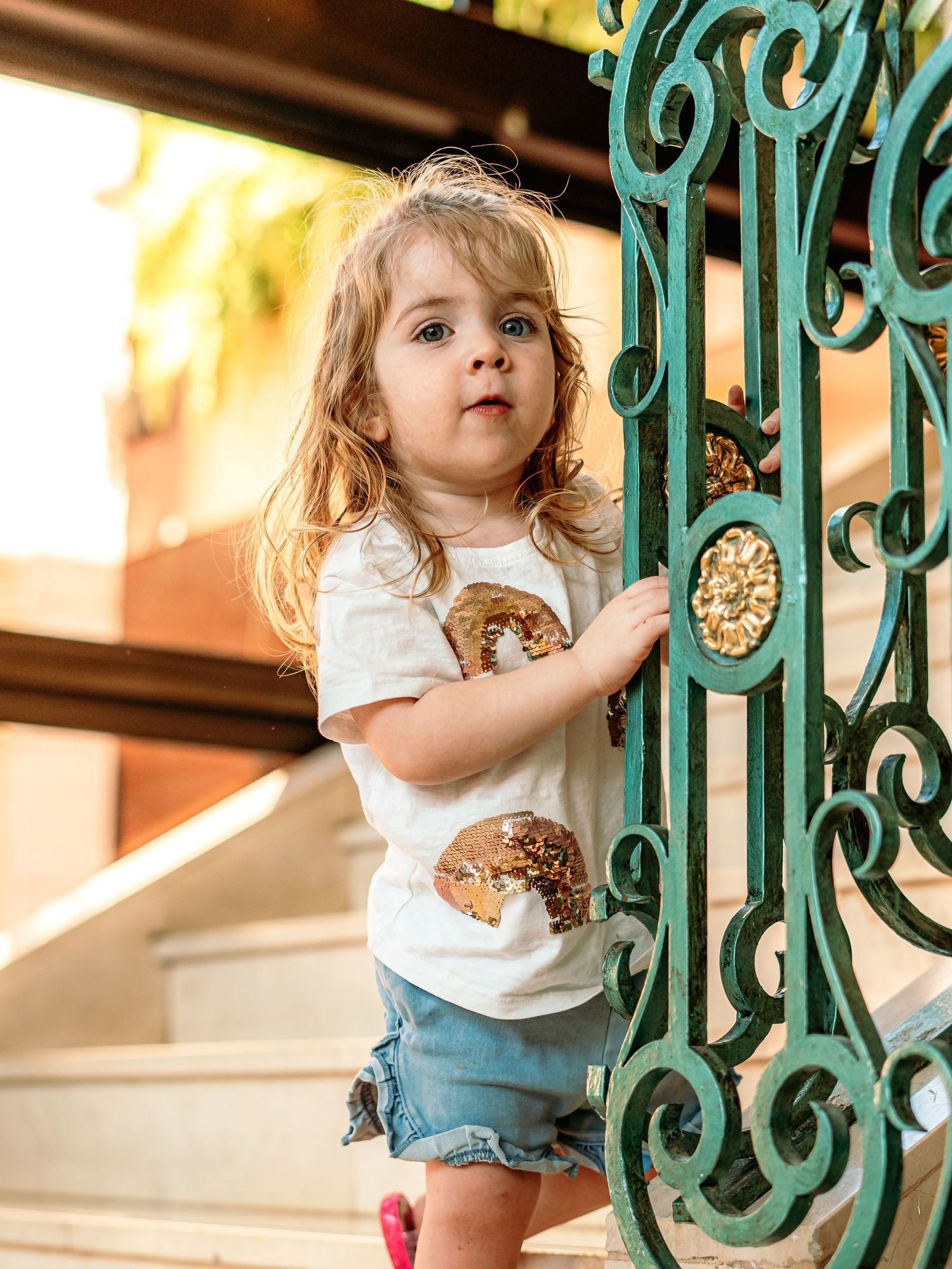 Blonde toddler girl in white sequin t-shirt holding decorative green iron gate.