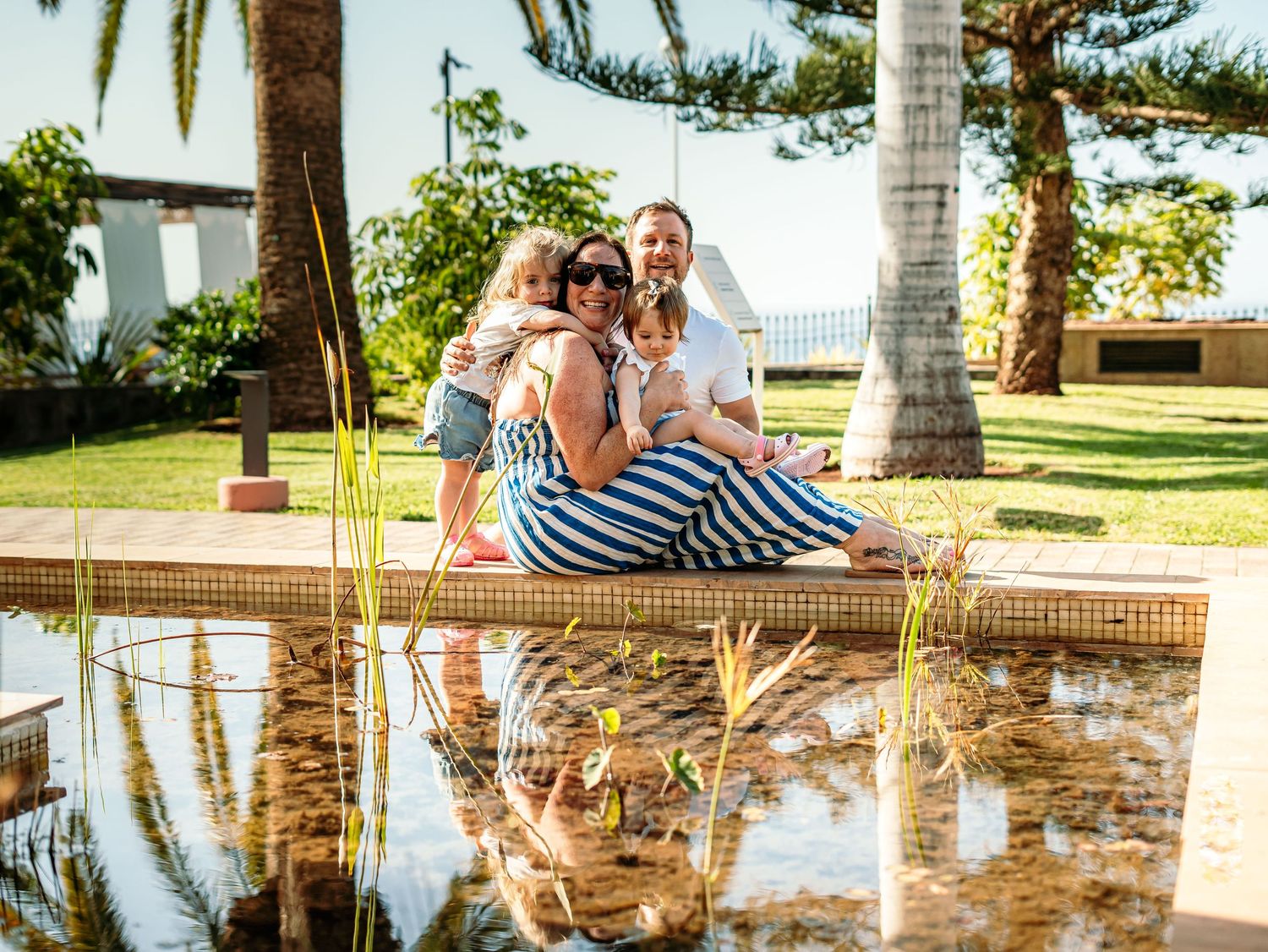 Family of four relaxing by a decorative pond in a tropical garden setting.