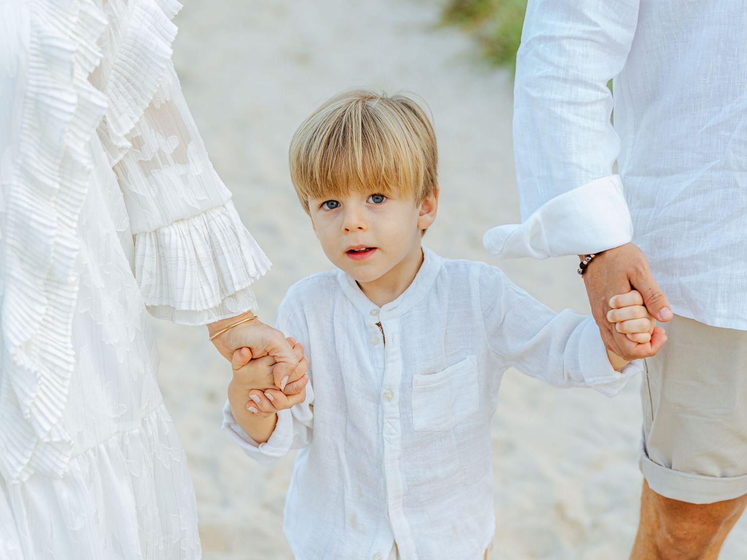 A young blonde boy in a white shirt holding hands with his parents on a sandy beach.