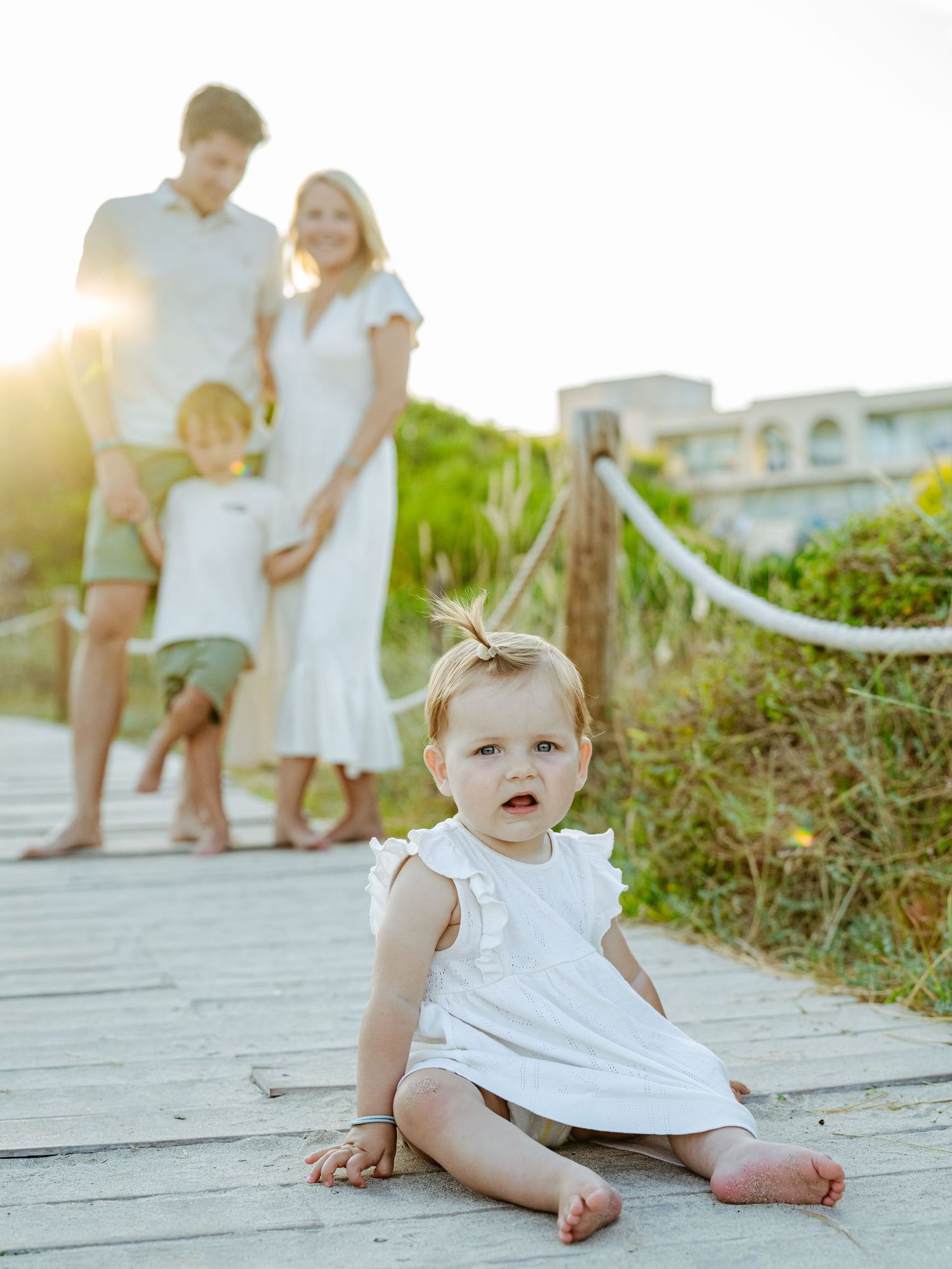 Baby girl in white dress sits on boardwalk while family poses behind at golden hour beach.