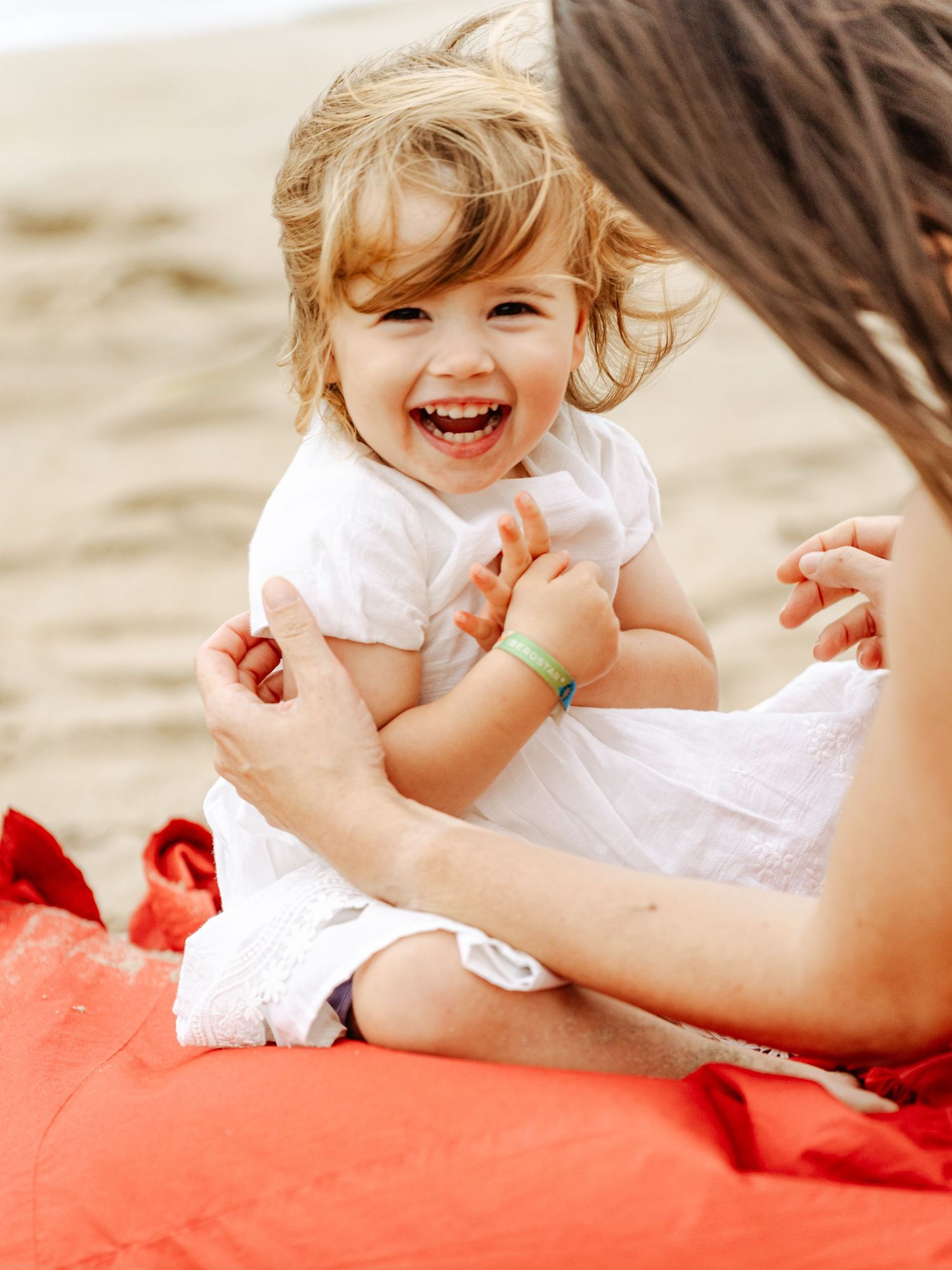 Laughing toddler girl in white dress sits on red blanket being tickled by her mother.