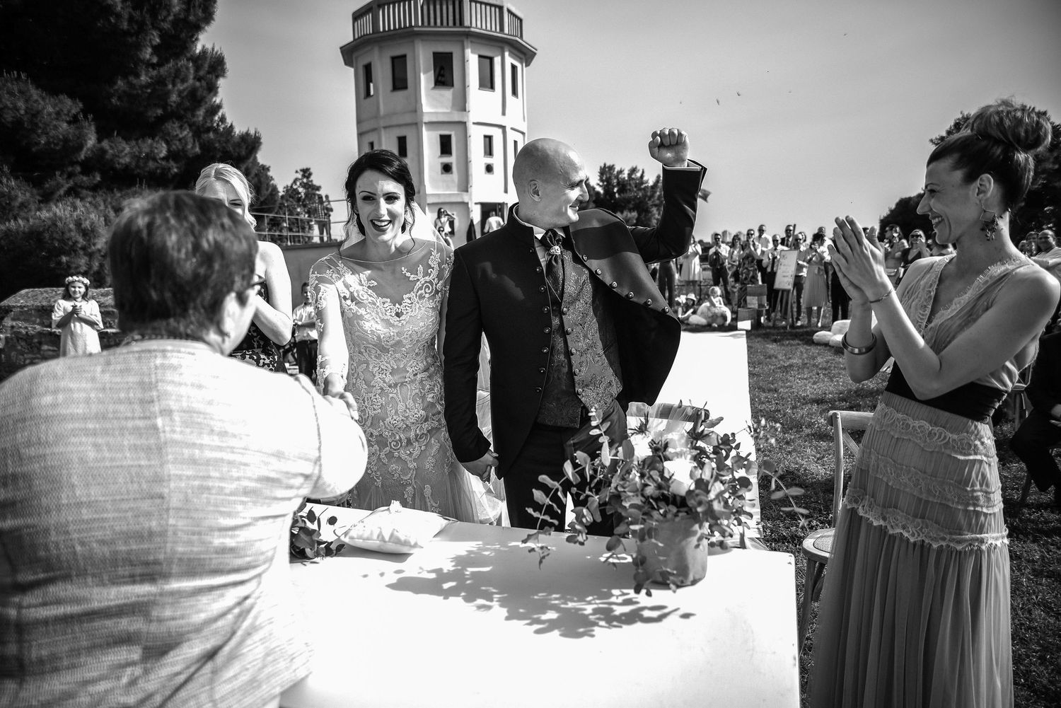 Black and white photo of newlyweds celebrating with guests near a historic tower after ceremony.