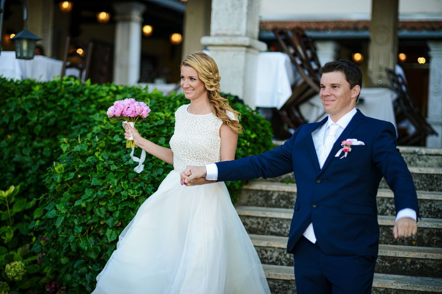 Smiling bride and groom walking down steps holding hands with pink bouquet at outdoor wedding.