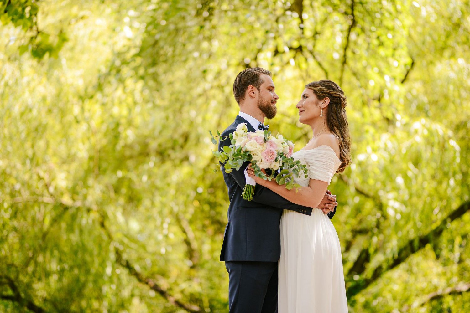 Bride and groom embrace romantically under lush green weeping willow trees holding a pastel bouquet.