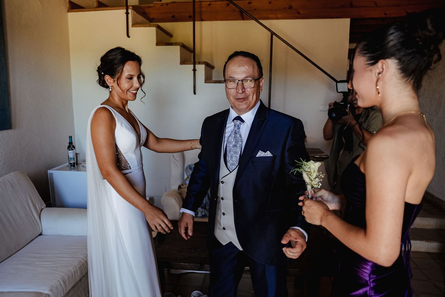 Bride in white gown smiling with father in navy suit as bridesmaid holds boutonniere nearby.