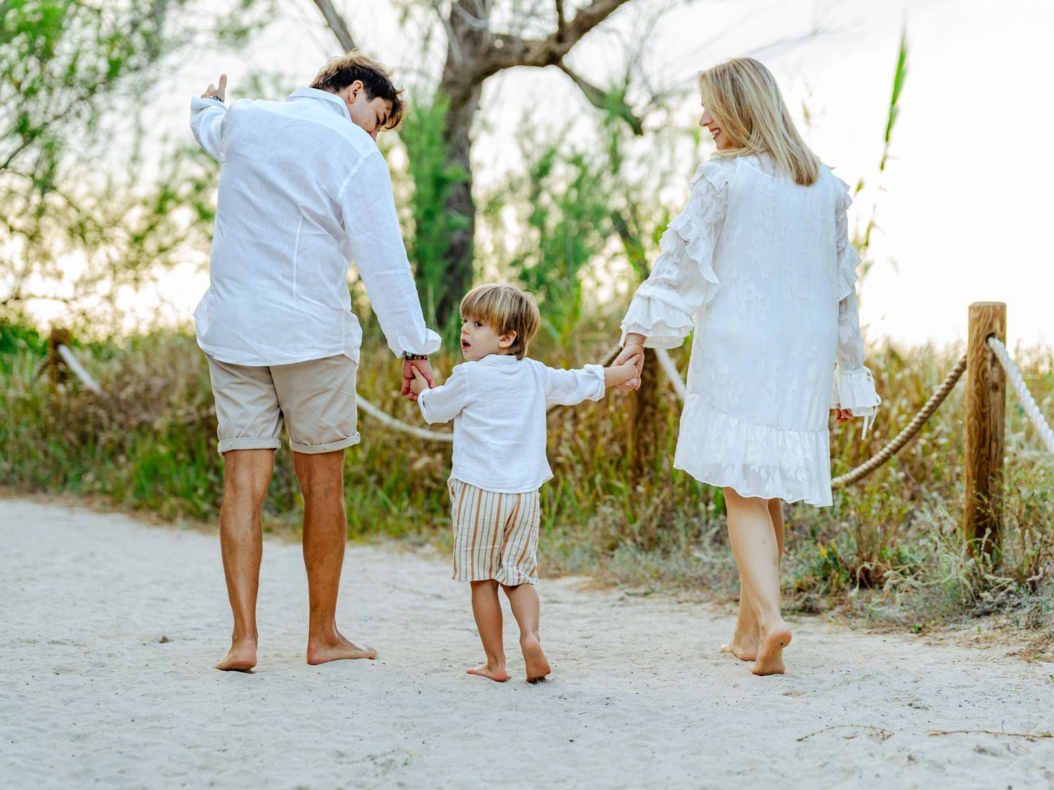 Family of three walking hand-in-hand on sandy beach path, all dressed in white, viewed from behind.