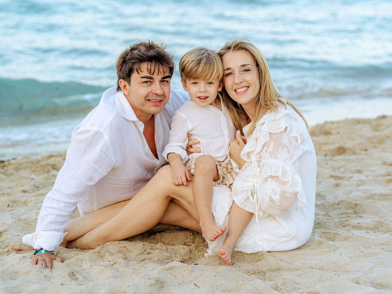 Happy young family sitting together on sandy beach smiling with ocean waves in background.