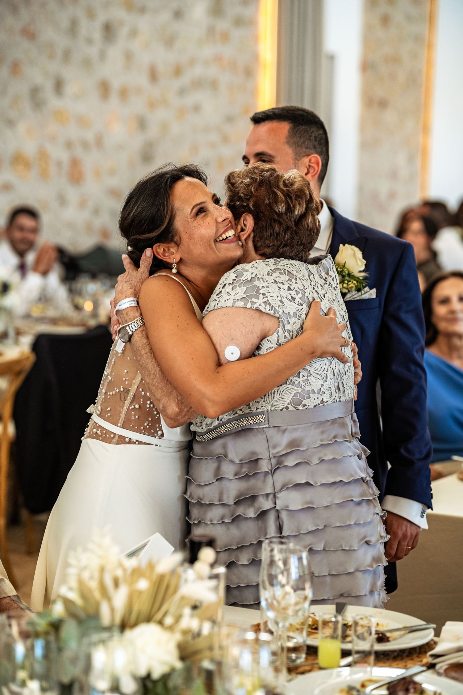 Bride shares a warm emotional hug with an older woman while groom stands close at the reception.