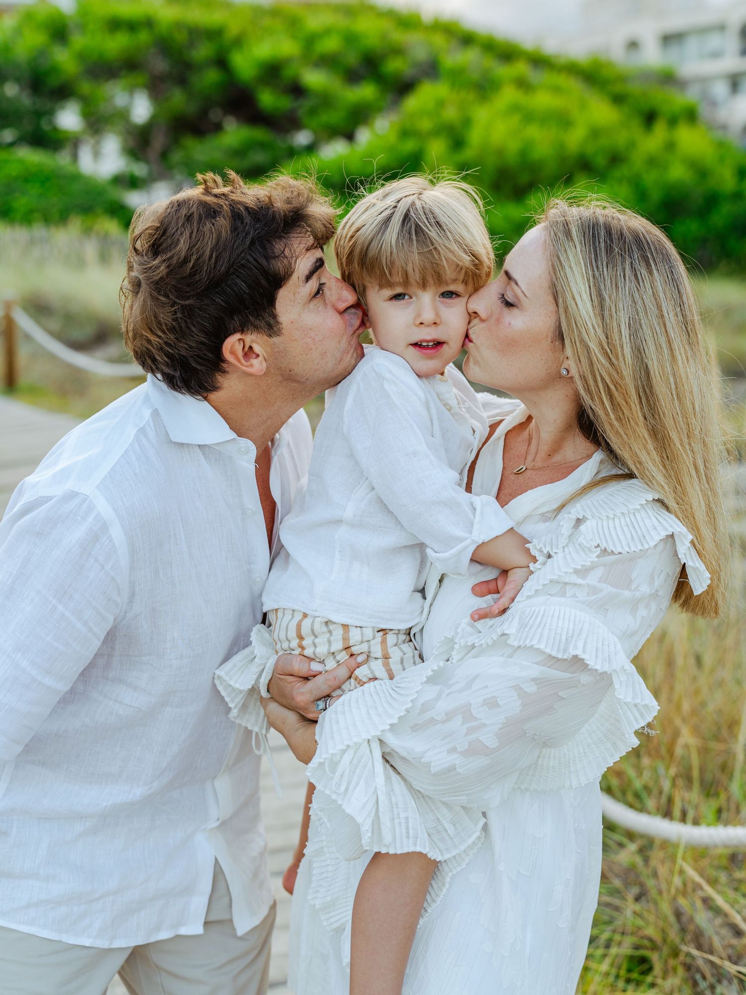 Parents in white outfits kissing their young blonde child on the cheeks outdoors.