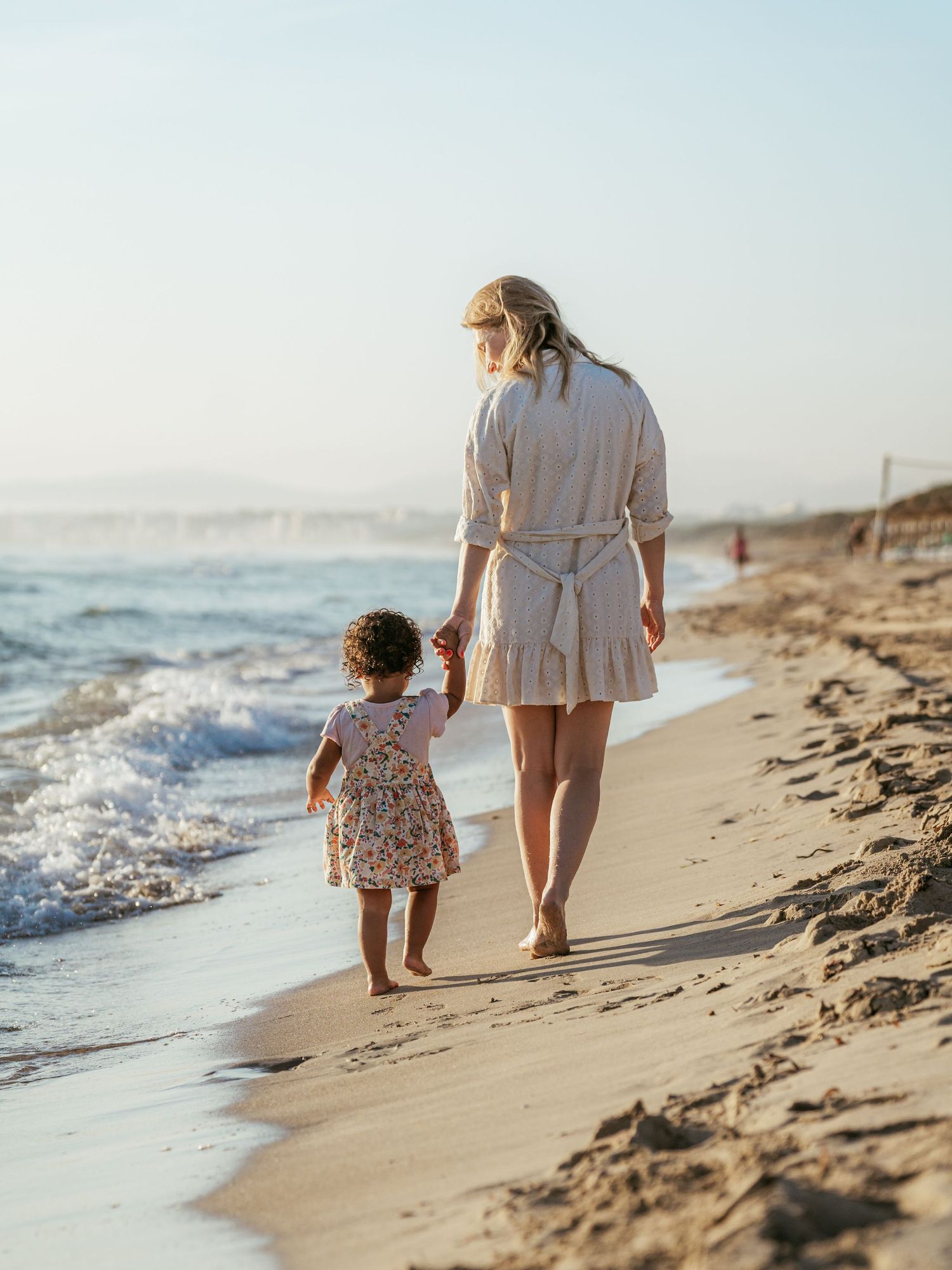 Mother and toddler walking hand in hand along a sunny sandy beach near ocean waves.
