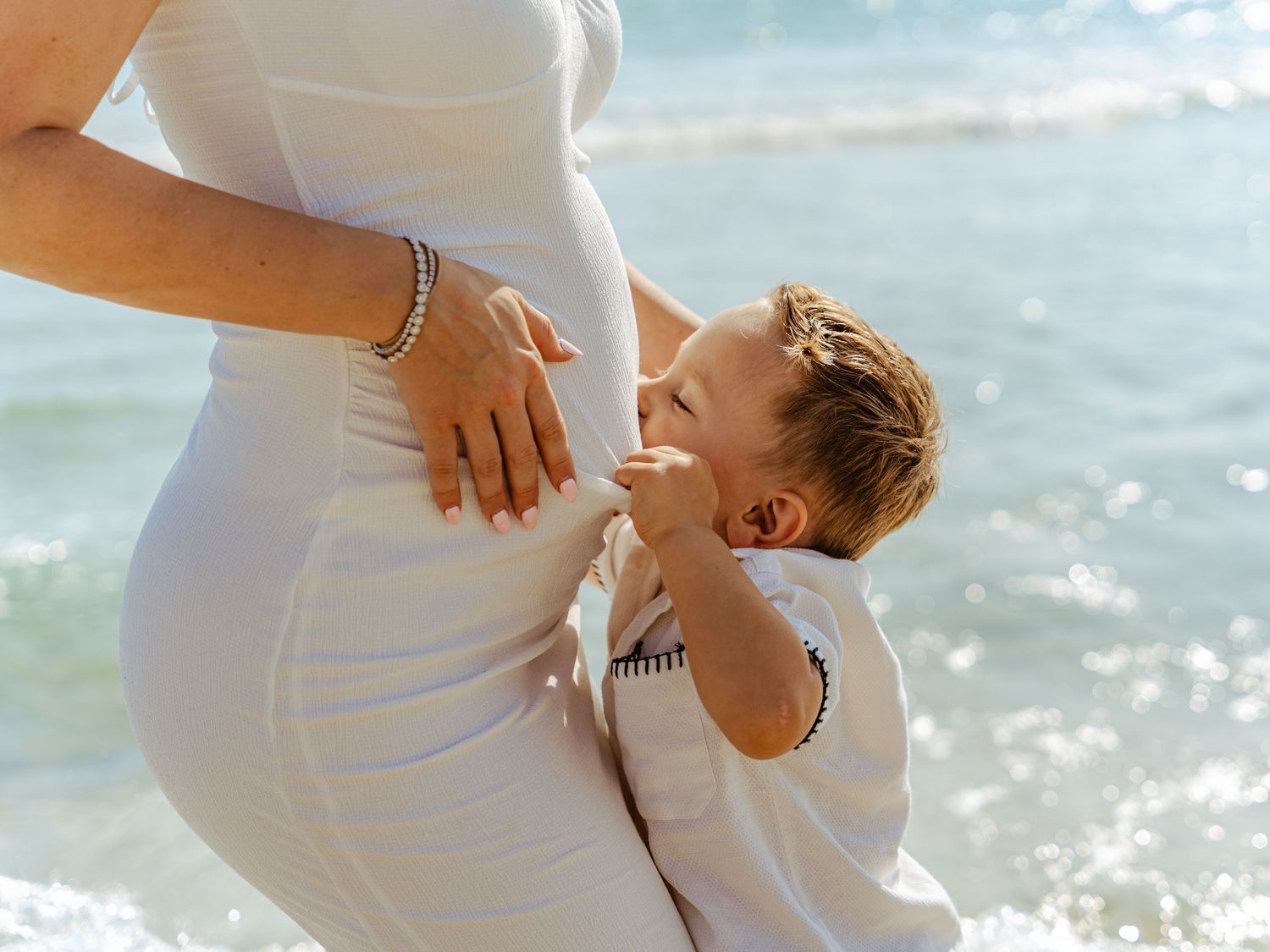 Young boy hugging pregnant mothers belly while standing together at the ocean shore.