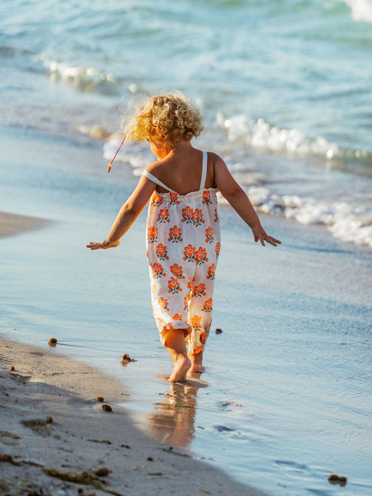 Curly-haired toddler in floral romper running barefoot along a shallow ocean shoreline at sunset.